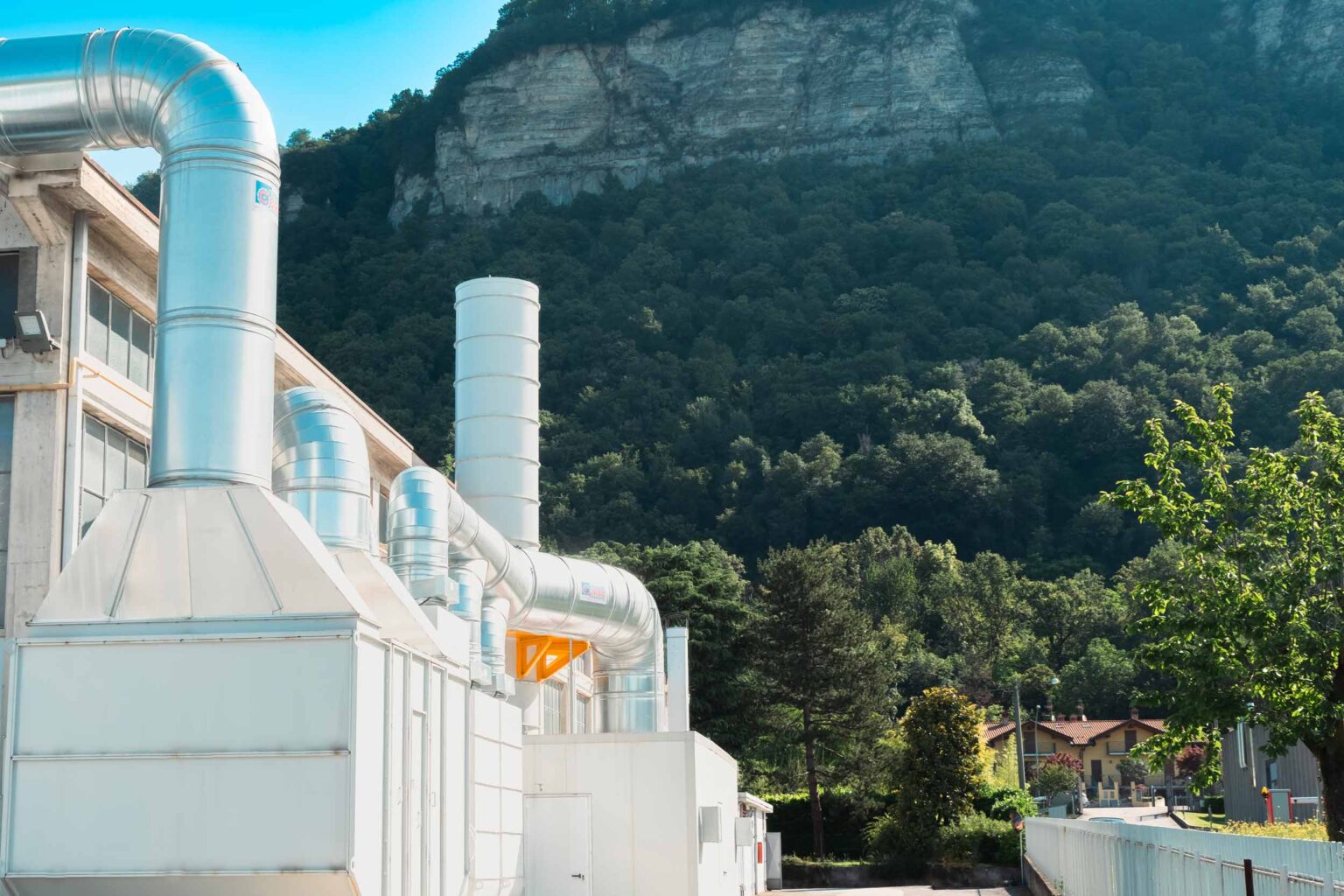 An exterior view of the Cisano Bergamasco plant, featuring large metallic ducts against a backdrop of lush, forested mountains.